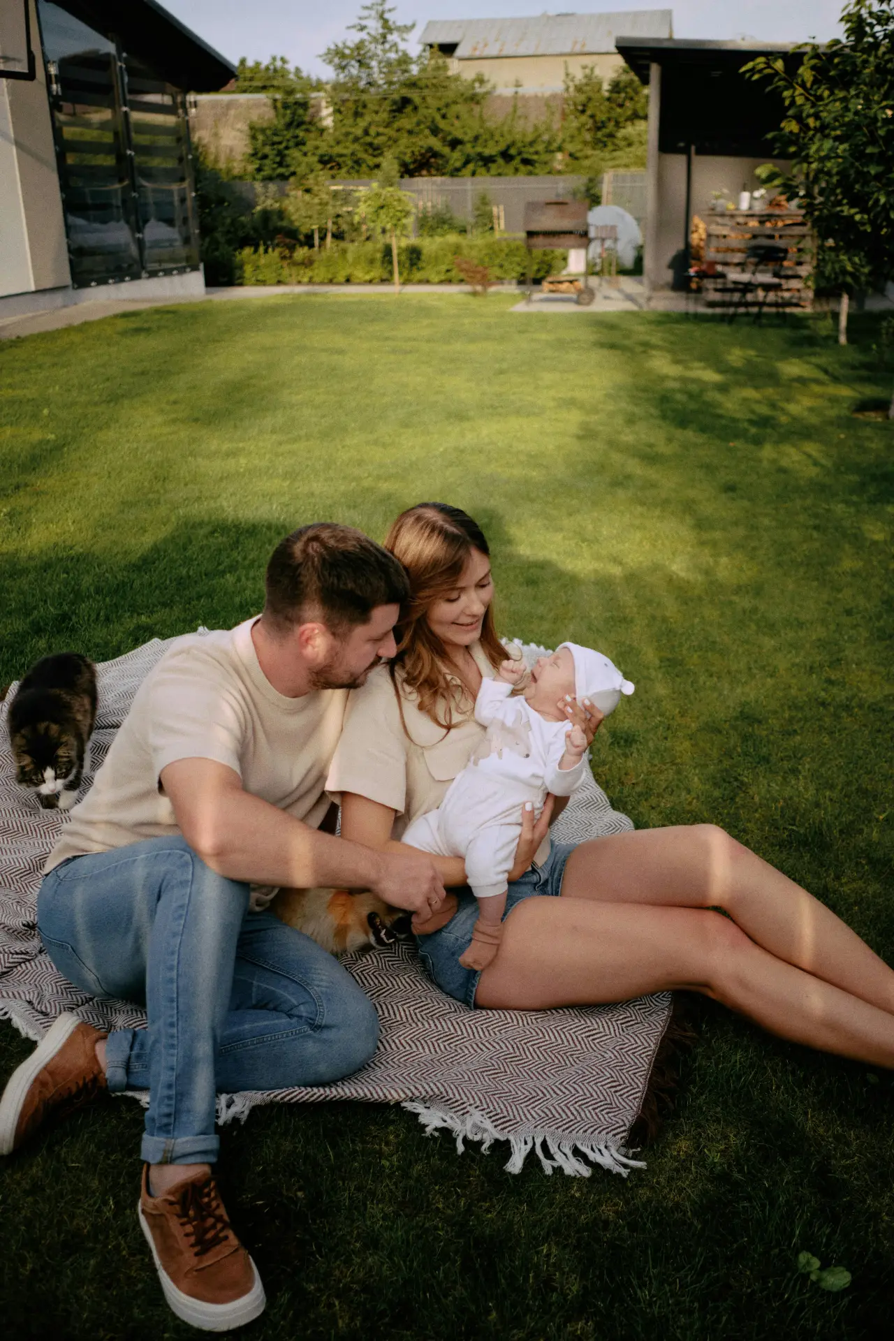 Ung familie med baby, katt og hund på et pledd i hagen en solrik kveld